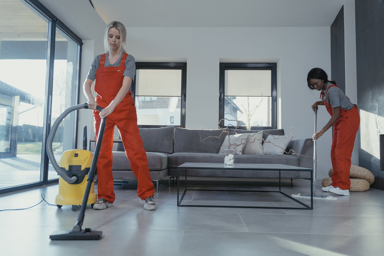 gallery-6 Two women in red overalls vacuuming and sweeping a modern living room.